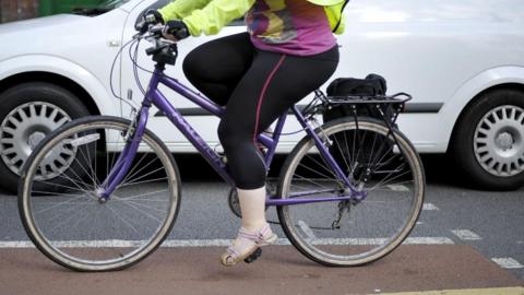 A close-up of the lower half of a woman on a purple bicycle on a road. She is wearing black leggings, sandals, a hi-vis jacket and gloves and there is a white van next to her on the road.