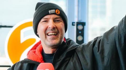 Paddy McGuinness smiling and waving in a black wolly hat and raincoat after finishing