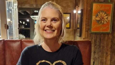 A woman in a black top sits in a restaurant on a brown leather bench. She has blond hair partially tied back and is smiling widely.