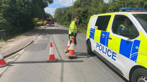 A man in high-visibility uniform puts down cones to close Cleat Hill, which is a residential road with trees either side. A police van also blocks the road. In the background there is a red fire engine with people stood next to it.