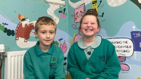 A blond boy in a green school jumper smiles, next to a girl who is grinning at the camera wearing a gingham dress under a green school cardigan in front of a colourful mural.