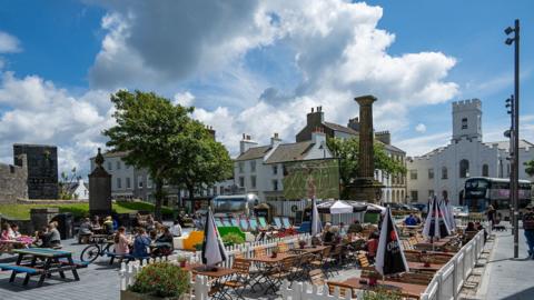 The square as it is pedestrianised over the summer, there are tables outside, some deck chairs, Wimbledon is being shown on a large screenn, you can see the castle in the background and people are sat around.