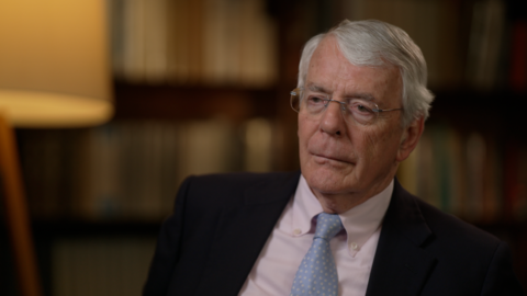 Sir John Major, wearing a dark suit, with a blue tie and white shirt, in a reflective mood
