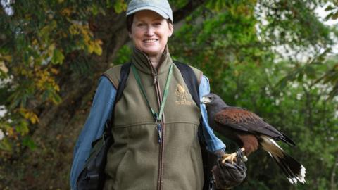Facing the camera, Candida is slim with short brown hair, she is wearing khaki all-weather clothing, and has a whistle on a lanyard