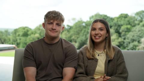 Fin and Sioned from Race Acoss the World series 5 - smiling towards the camera and sat on a sofa with trees behind them.