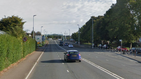 Google image shows Bridge Road near Exeter. Cars are driving along either side of the carriageway. Trees, bushes and lamp-posts line either side of the road. A footbridge can be seen in the distance which goes over the top of the road.