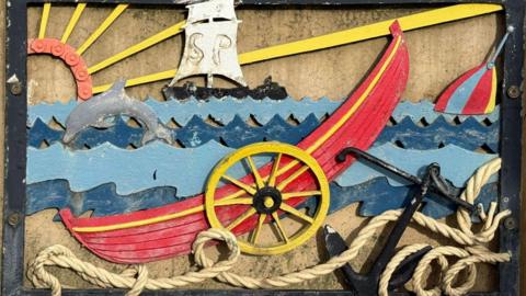A wrought iron depiction of a red ship on a rough sea with an anchor in the bottom left of the postcard. A dolphin is leaping from the waves in the background. It has been painted in bright colours but which are fading. 