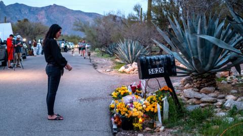 A woman wearing a dark top and jeans stands in quiet contemplation by a flower memorial outside Nancy Guthrie's home
