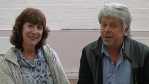 A woman and a man smile at the camera. The woman has shoulde-rlength brwn hair and wears a floral jumper and grey zip-up. The man wears a blue shirt and grey and black zip-up jacket.