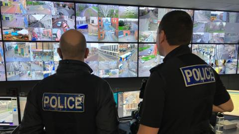 The image shows two police officers standing in front of a large array of surveillance monitors. The officers are wearing branded police uniforms. The monitors display various street views from different locations.