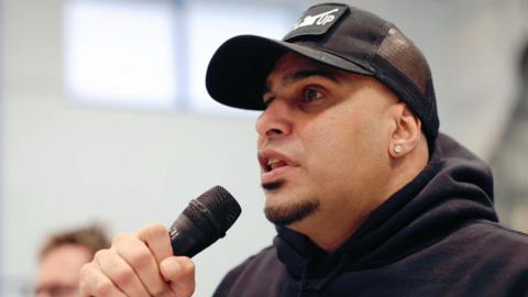 An image of Marcellus Baz holding a microphone whilst wearing a cap