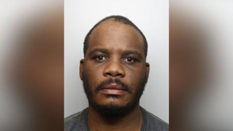 Kennedy Jimmy is looking at the camera in a police custody shot. He has very short black hair and a black beard and moustache. He is wearing a green T-shirt.