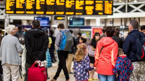 Adults and children look at train departure boards at a train station. Some are carrying luggage.