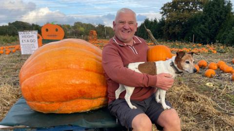 A man sits on a blue pallet holding a giant pumpkin. The man is hugging a small white and brown dog which sits on his lap and a regular sized pumpkin in his other hand. The min is wearing a maroon long-sleeved top and navy shorts. Behind him is a sign which reads 'Guess the weight. £1 a go' The field they are in is filled with pumpkins laying on the soil. Trees and cloudy sky can be seen behind.