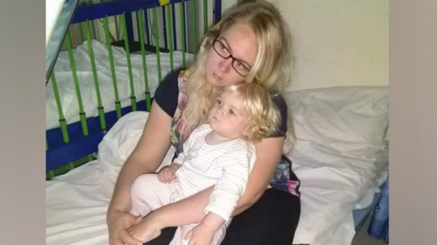 A woman with blonde hair and glasses, looks worried as she cradles a young toddler, with blonde hair, wearing a white baby grow. Both are sitting on a hospital bed