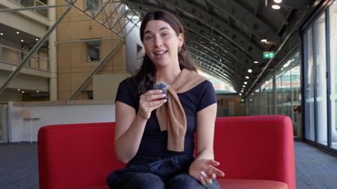 Young woman with brown hair holds a microphone while sitting on a red couch