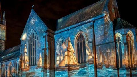 A church lit up as part of a light installation. Orange ships can be seen on blue water as part of the installation. The sky is dark.