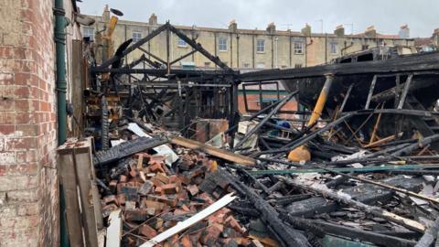One of Underfall Yard's buildings is shown completely gutted by the fire. Just the frame of the building remains, with the roof and even exterior walls completely caved in. Piles of rubble fill the foreground.