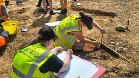 Two young women in flourescent vests using a tape measure on muddy ground next to stones