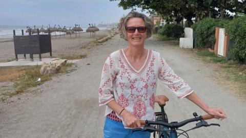 A woman in a white top and blue trousers holds a bicycle by a beach. 