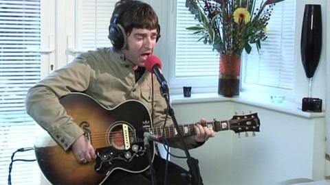 Noel Gallagher, with mid-length dark hair and wearing a beige denim jacket and black jeans, sits on a chair and plays a brown guitar in front of the windows of a conservatory and a vase of flowers