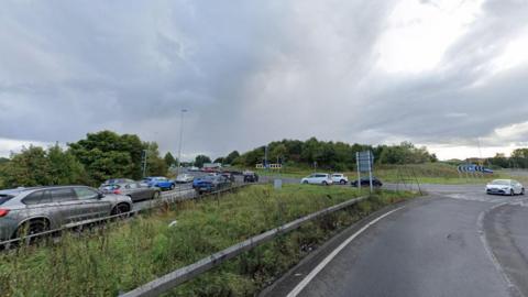 Moor Farm roundabout on the A19. The roundabout is covered by trees and grass. A queue of traffic is waiting to join the roundabout on the left.