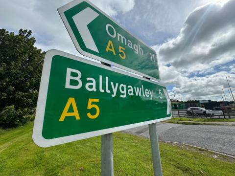 Green road signs with 'Omagh A5' and 'Ballygawley' A5 on them. Trees and green grass in the background. The sky is grey with clouds and a car is parked in a carpark behind the signs. 