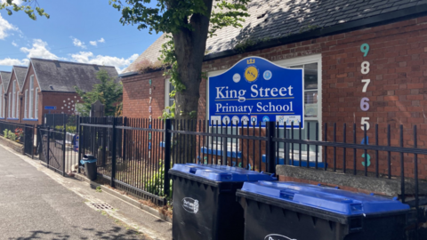 A picture of the outside of a school with a blue sign with the words King Street Primary School in the foreground.