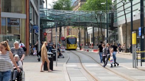 Manchester city centre on a busy day, with pedestrians seen passing over tramway close the the Arndale shopping centre, with a yellow bee network dram seen in the distance, and a public walkway connecting two buildings pictured above.
