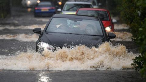 A black car, at the head of a queue of cars, drives through deep flood water