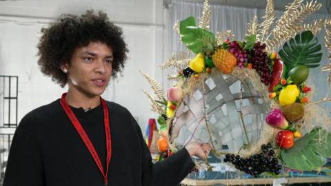 A teenage boy with curly black hair, wearing a black jumper and red lanyard, holds a large headpiece covered in fruit and gold leaves.