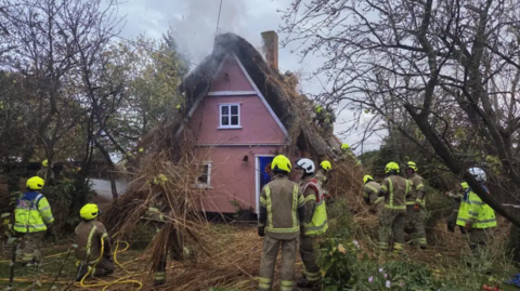 Several firefighters standing around a pink thatched cottage which has smoke coming out of its roof. The building is set amongst a woodland area 