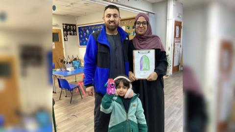 Akeel, Hannaa and Sara in Al Ashraf Primary School. Sara is wearing a warm coat, pink earmuffs and has a hand puppet. Hannaa is holding a drawing done by Sara.