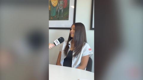 A young girl with brown hair sat in a chair. She's facing a reporter who is holding a mic to her face.