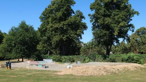 Grass park with skate park featuring slopes to the left and a higher platform on the right. There is graffiti on the ramps. There are two people standing at the edge of the skate park. There are trees and a hedge beyond the skate park.