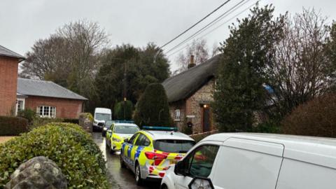 Lane with homes either side and numerous vehicles including police cars parked along.