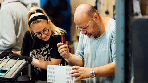 A man using a screwdriver on a machine while a woman watches 