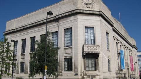 A large white brick building covers a corner of a street. There is a balcony, large windows and colourful banners hanging from one wall