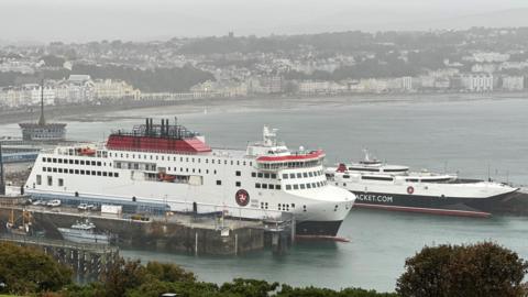 The Manxman and Manannan ferries moored in Douglas Harbour. The are both in the Steam Packet colours of white, red and black. Douglas bay and promenade are in the background with a grey sky above.