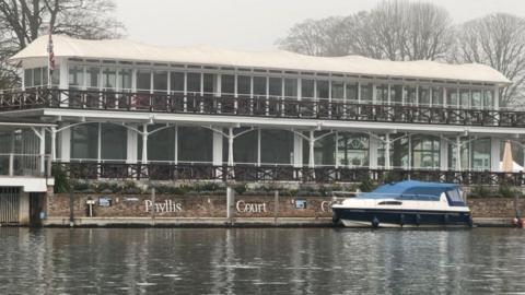 Phyllis Court pavillion is right next to the river Thames. It is a glazed building with white supports. In the foreground is the river with a boat moored alongside the building. There is a large white sign reading Phyllis Court on a brick wall in front of the building.