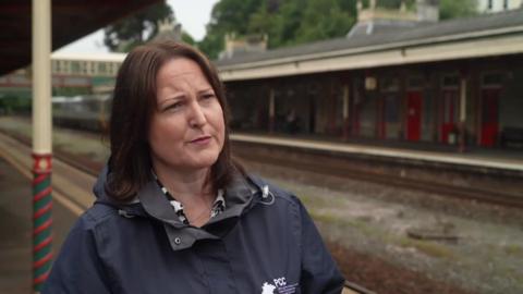 A picture of Alison Hernandez standing at a train station. She is wearing a black coat and has brown hair.