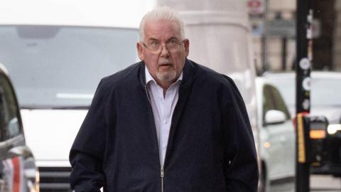 Andy Osborn arriving at court. He has short white hair and a goatee beard. He is wearing a dark zip up top and a light shirt underneath.