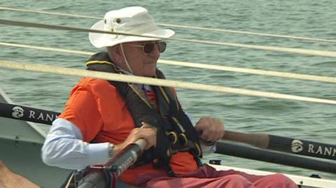 A man wearing orange sitting in a boat holding two oars. He is wearing sunglasses and a white hat.