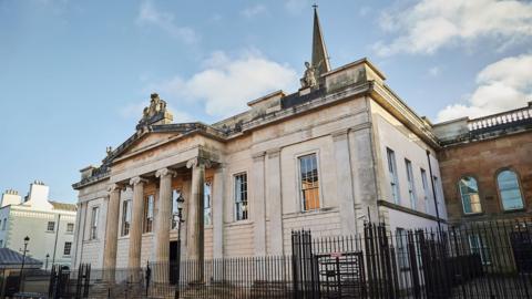 Crown court in Londonderry pictured with a black fence around the exterior. It is a stone building.