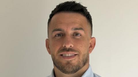 A headshot of Nicky Kingsnorth, who has black hair, short at the sides, and a close-cropped black beard. He is smiling. His light blue shirt collar can just be seen.
