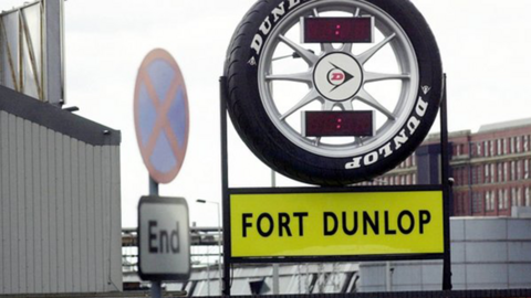 The sign for Fort Dunlop, comprising a Dunlop tyre and yellow sign with black writing underneath it. The three-storey former factory building can be seen behind it.