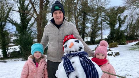 A man in a navy hat with a green crest and grey zip up top stands with two little girls in pink coats and a snow man in front of them