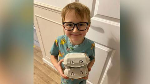 A boy smiling straight at the camera while holding two egg boxes. He is wearing blue pyjamas and glasses.