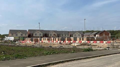 A building site in the foreground with piles of bricks behind small plastic barriers. Rows of new detached homes can be seen further back.