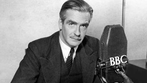 Black and white. Anthony Eden sitting in front of a microphone with "鶹Լ" written on it.  He is wearing a suit and tie and looking down the lens.